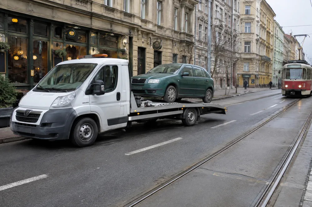Bílá odtahovka do 3,5 t nakládá zelenou Škodu Fabii v žižkovské ulici vedle tramvajové trati